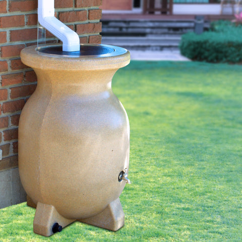 A stone looking rain barrel sits next to the brick wall of a home. The downspout is located directly above the rain barrel allowing the water to pour into it. Next to the rain barrel is the trunk of a tree.