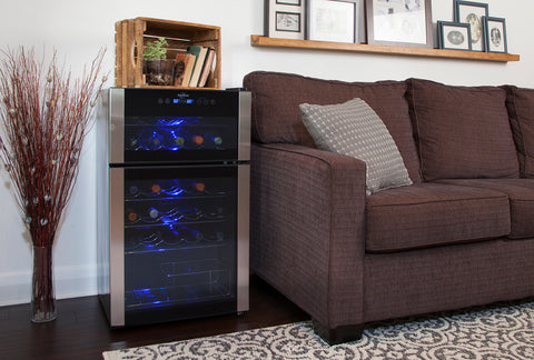 A stainless steal wine fridge sits on the floor next to a grey couch. Above the couch are picture frames. On top of the wine fridge is a wooden basket with books and a plant in it. Beside the wine fridge is a vase with decorative sticks in it.