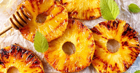 Photo of grilled sliced pineapple on parchment paper with wooden honey drizzle stick and mint leaves