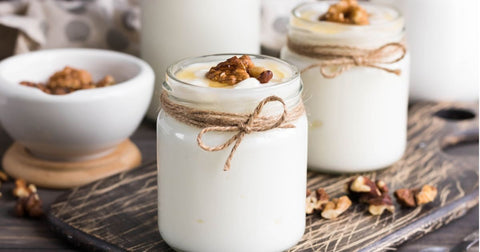 Two small jars of plain yogurt topped with chopped nuts and honey and a bowl of nuts on a dark gray wooden surface