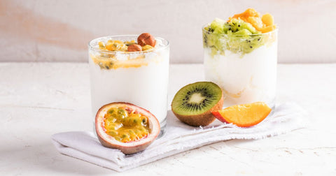 Photo of two glass jars of plain yogurt topped with fresh fruit and nuts on a white painted table with slices of passionfruit, kiwifruit, and peach beside them