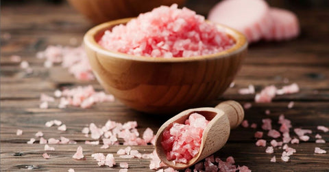 Picture of a wooden bowl and wooden scoop both filled with coarse pink salt crystals with more salt sprinkled around then on a wooden surface