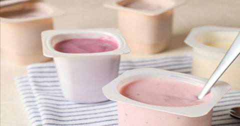 Photo of 5 opened single-serving yogurt cups, one with a spoon in it, sitting on a tabletop and a folded blue and white striped napkin