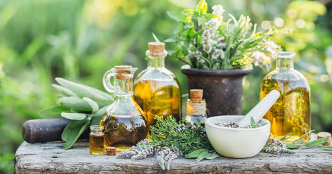 Picture of bunches of fresh herbs (sage, basil, lavender), glass jars of olive oil, and a mortar and pestle on a rustic wooden bench