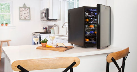 Photo of a 20 bottle single zone thermoelectric wine cooler open and filled with bottles of red and white wine on a white kitchen peninsula. There is a wooden cutting board with cheese wedges and two glasses of white wine beside it