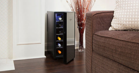 Photo of 12 bottle, single-zone thermoelectric wine fridge, open with bottles of wine inside, on a dark wooden floor with white painted wainscotting behind and a glass vase of dogwood branches and a light brown sofa to the right 