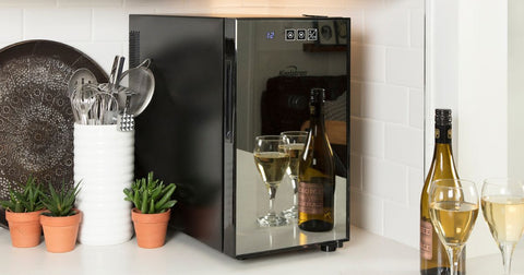 Closeup image of a small thermoelectric wine cooler on a white countertop with a white tile backsplash behind and a decorative plate, three small plants, and a white container of metal kitchen utensils beside it. The glass door of the wine fridge is mirrored and reflecting a bottle and two glasses of white wine placed in front.