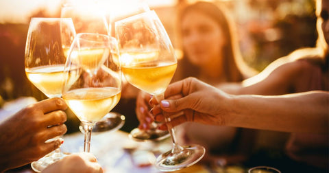 Closeup image of hands clinking glasses of white wine together with blurred faces in the background