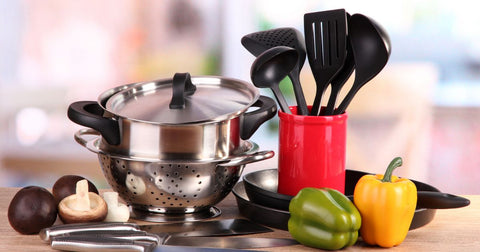 Photo of kitchen items including a strainer, cooking pot with lid, knife, and black spoons and spatulas in a red container, as well as several vegetables arranged on a counter