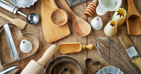 Photo of kitchen items including wooden mixing spoons, flippers, rolling pins, measuring cups, icing tips, graters, whisk, and cookie cutters, arranged on a wooden counter and viewed from above