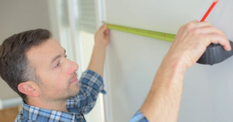 Picture of a person with light skin and short dark hair and facial stubble wearing a blue plaid shirt measuring a wall with a yellow measuring tape