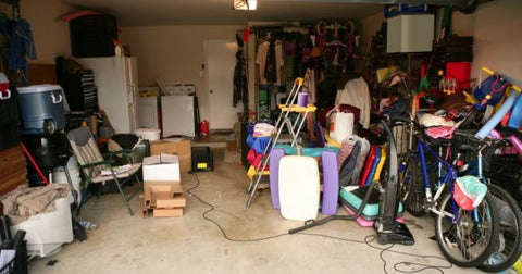 Picture of a very cluttered garage filled with items like a ladder bikes, pool toys, a children's picnic table, cardboard boxes, etc.