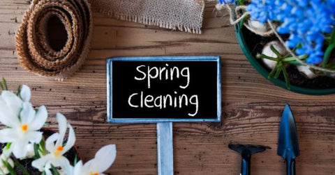 Picture of a small chalkboard with text reading spring cleaning lying on a wooden board surrounded by blue and white flowers, burlap fabric, and gardening tools