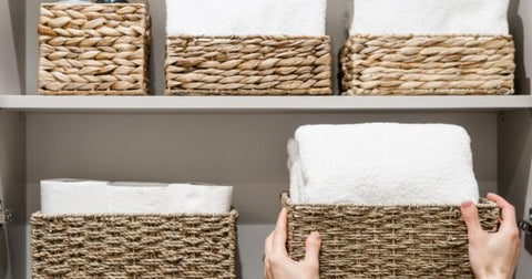 Closeup image of a person's hands placing a brown woven basket filled with white towels on a shelf that contains four other similar baskets