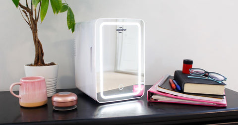 Photo of mirrored cosmetics fridge on a black desk with ring light turned on and school supplies beside it