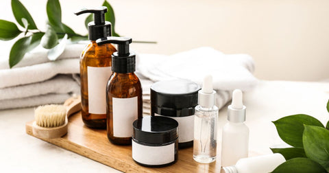 Several bottles and jars of lotions and serums on a wooden tray with white towels and green leaves in the background