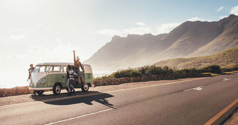 Picture of three people leaning out of a white and green VW microbus parked at the side of a remote highway with mountains in the background