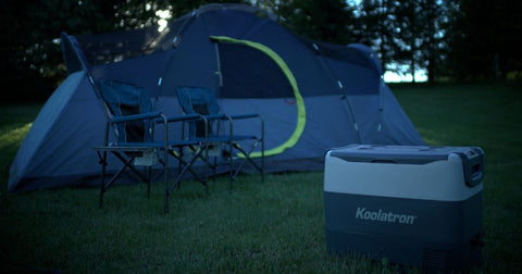 Picture of a campsite at night with a Koolatron SmartKool on the grass in front of two folding camping chairs and a dome tent