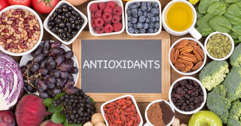 An assortment of antioxidant-containing fruits, vegetables, nuts, and berries, some in bowls or cups, is pictured from above. There is a small chalkboard in the middle with the word ‘antioxidants’ written on it