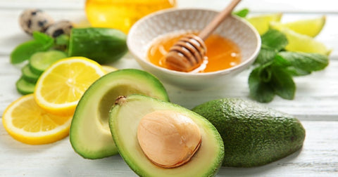 Lifestyle image of lemon slices and avocado halves arranged around a small white bowl of honey