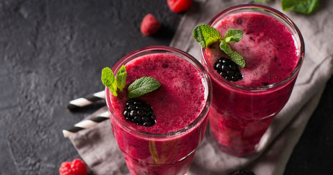 Lifestyle image of two glasses of vibrant purple red smoothie topped with mint leaves and a blackberry on a dark gray table