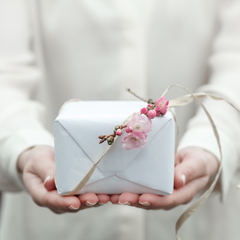 hands holding out a present wrapped in white with a pink flower