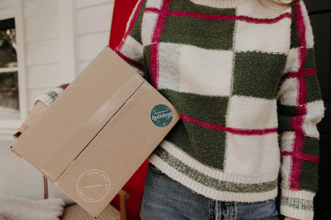 green and pink plaid sweater, holding shipper with a sticker reading "don't open until the holidays"