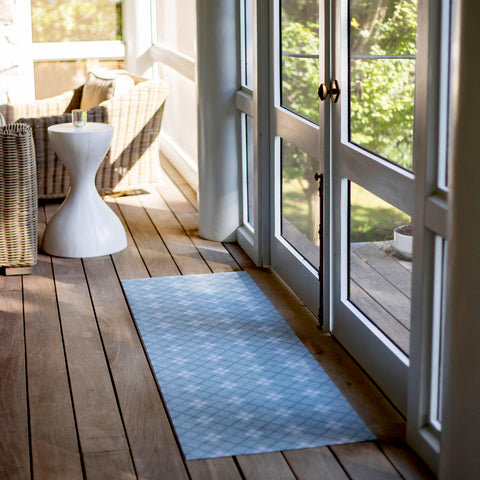 A bright and airy interior space with a light blue patterned mat at double glass doors. The room has a cozy sitting area with wicker furniture and a small white side table, creating a serene atmosphere with natural light streaming in.