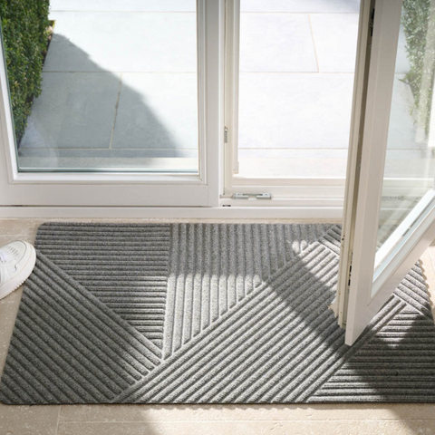 A grey WaterHog® door mat with a geometric pattern just inside an open glass door. The mat effectively traps dirt and moisture, helping to keep the light-coloured tiled floor clean and dry, with a glimpse of green visible outside the entrance.