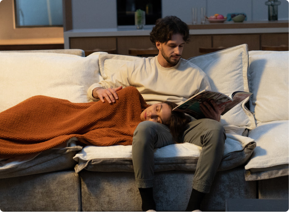 a man is reading on the sectional sofa with a woman sleeping on his legs