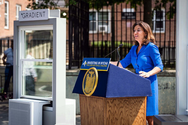 A woman in a blue dress speaks at a podium next to a window with a Gradient air conditioner