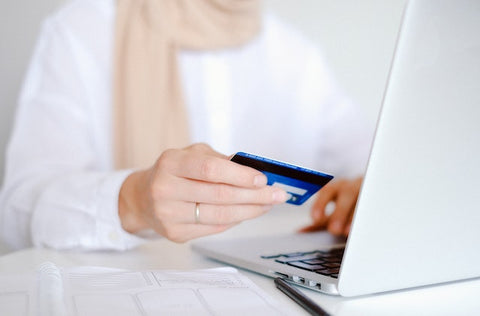 A person wearing white clothes sitting in front of a laptop and holding a credit card