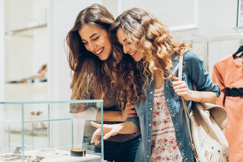 Two women are shopping and browsing a jewelry display case. Both are smiling and one of the women is pointing at the case.