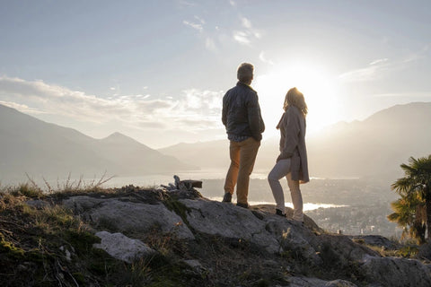 An older couple is standing on a rock overlooking a natural landscape. They are looking at a lake in the distance with the sun backlighting them.