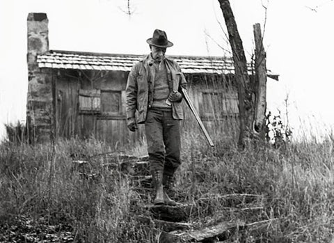 Black and white photograph of Aldo Leopold carrying shotgun down a scrubby hill with small building in background.
