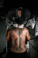 A person with a darker skin getting a tattoo on his back, photographed from the top