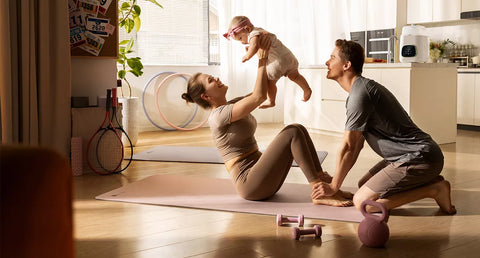 A mother is sitting on an exercise mat in the living room, holding her baby while doing leg exercises with her partner pressing down on her feet for resistance.