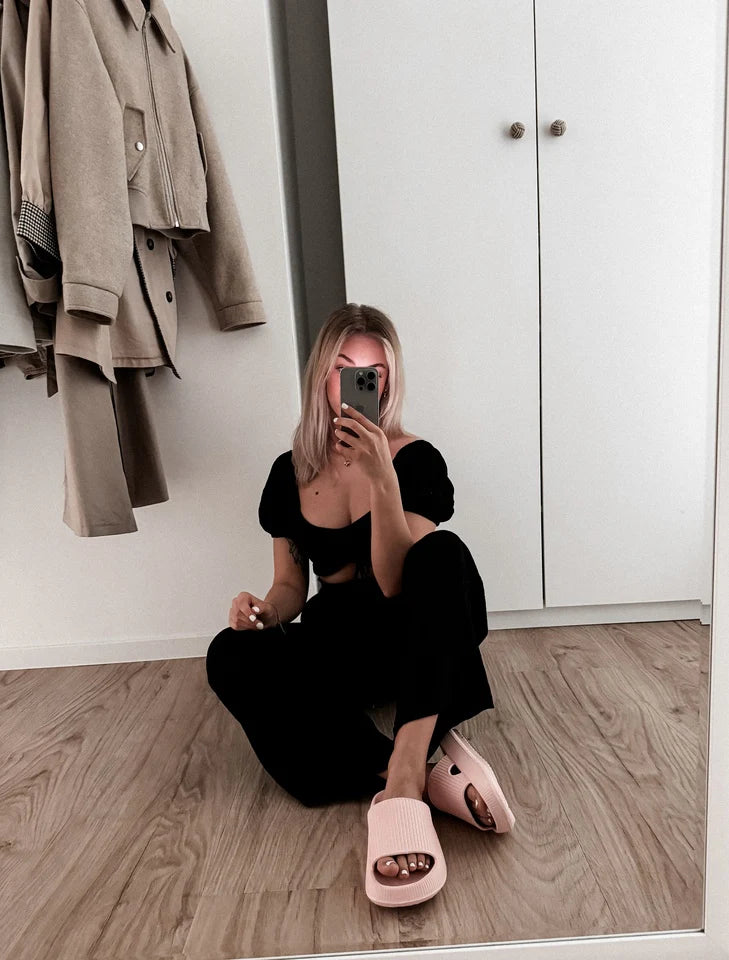 Persona tomándose un selfie en el espejo en una habitación elegante, con ropa negra y zapatillas rosas.