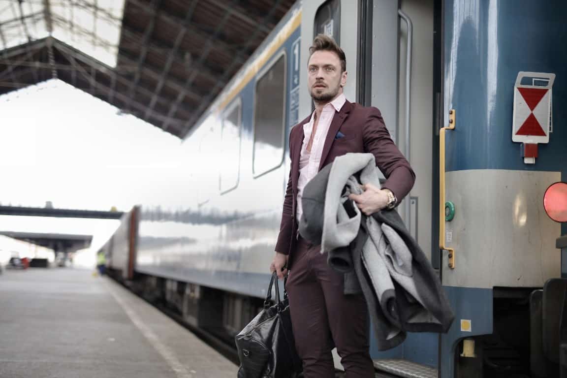 Tinute elegante de barbati pentru iarna - barbat in costum visiniu, cu geanta, in fata unui tren