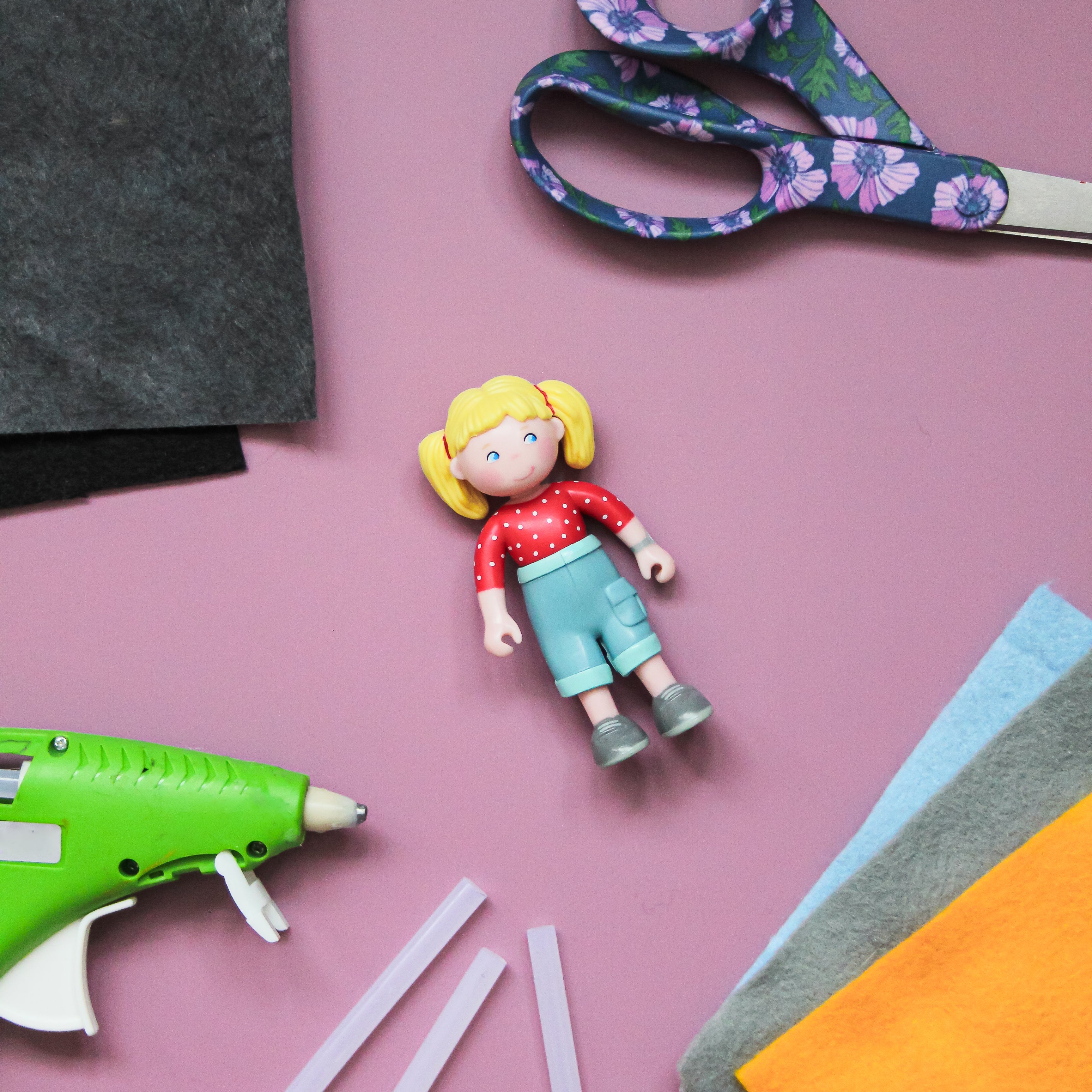 Une poupée Little Friends se trouve au milieu de feutre, d'un bâton de colle et de ciseaux sur un fond mauve clair.