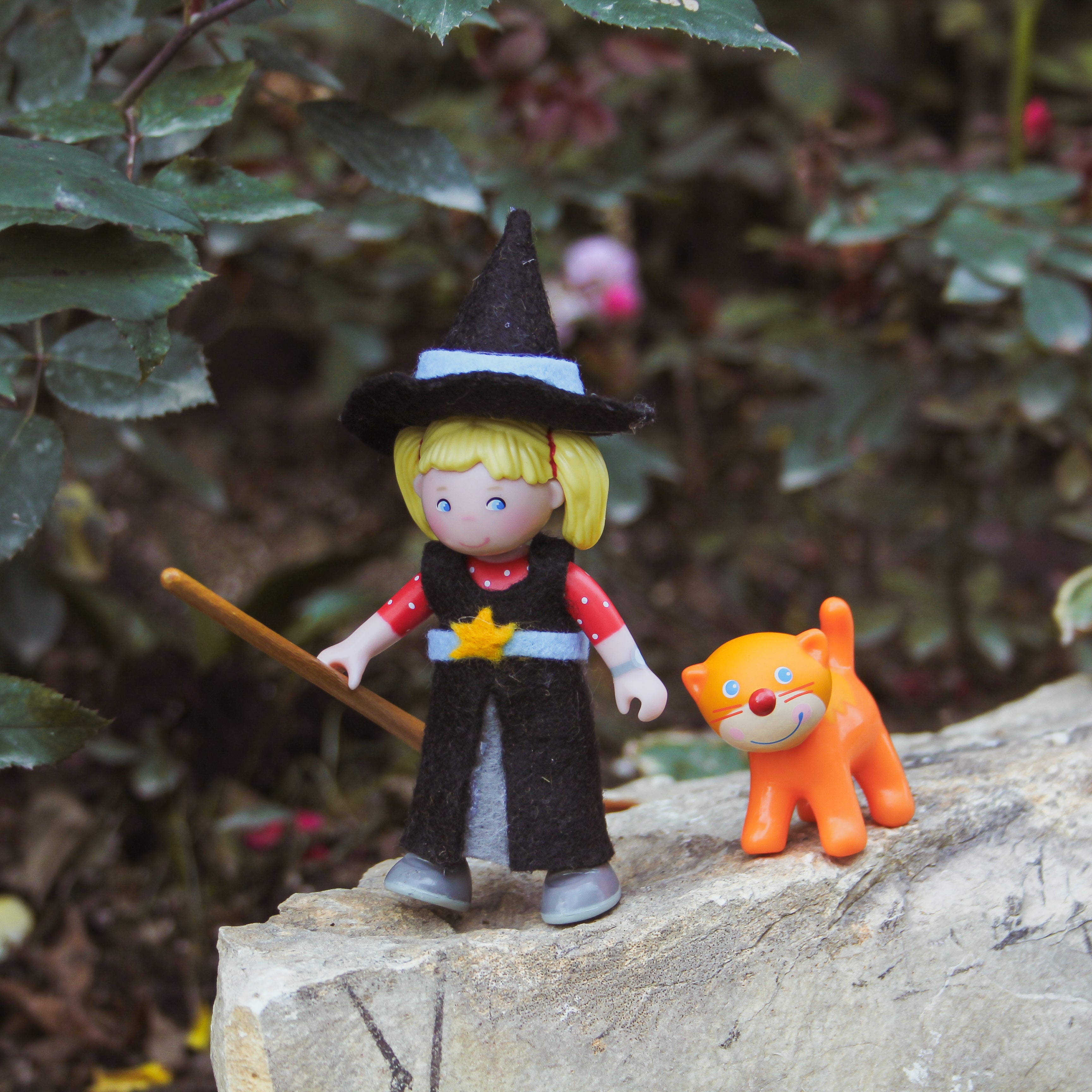 A Little Friends doll in a witch costume with an orange cat stands on a rock.
