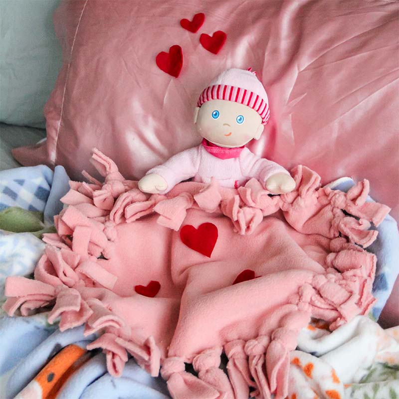 A Luisa doll is snuggled under a pink blanket.