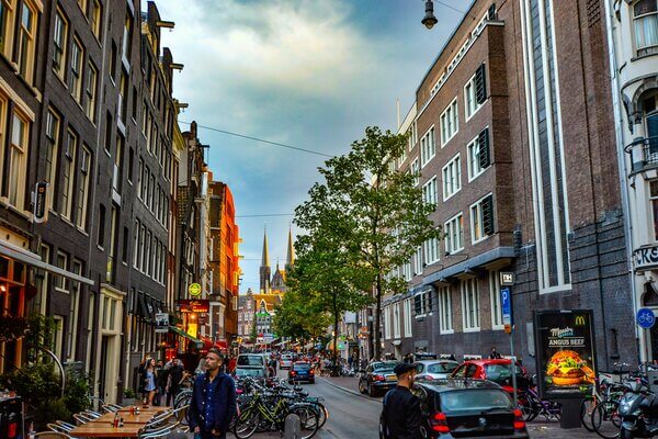 Druk gedeelte in Amsterdam, op de weg rijden auto’s en langs de straten staan veel fietsen geparkeerd en wandelen mensen.