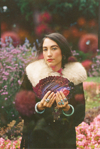 Alejandra Luisa León standing in a field of flowers holding the Stars Divine astrology deck cards fanned out in her hands