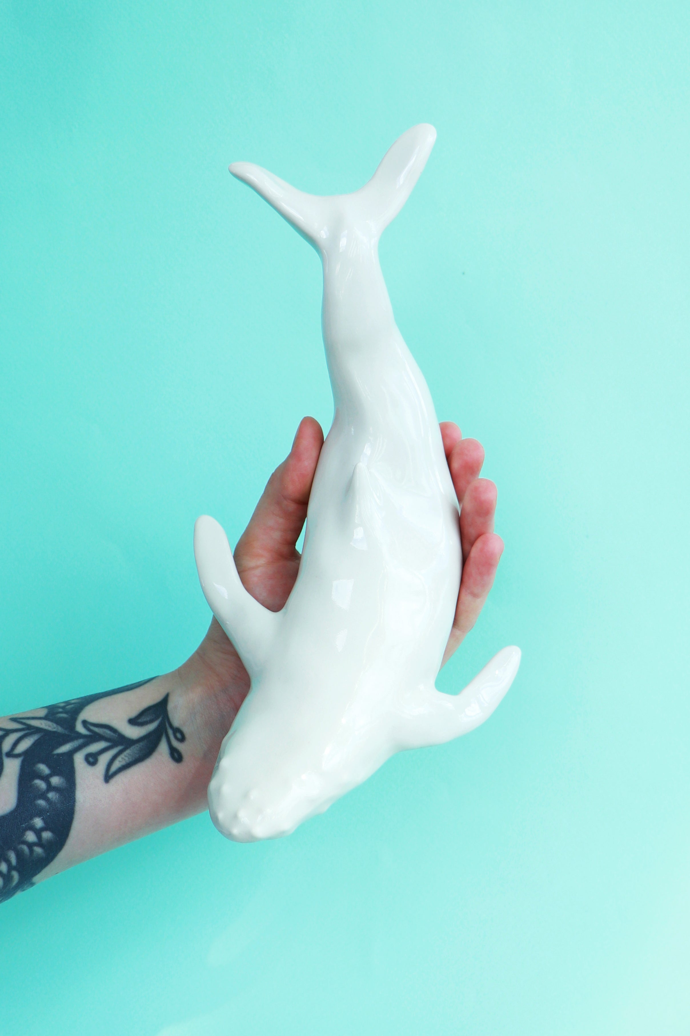 Ceramic humpback whale against a turquoise background. A tattooed arm is holding the whale.
