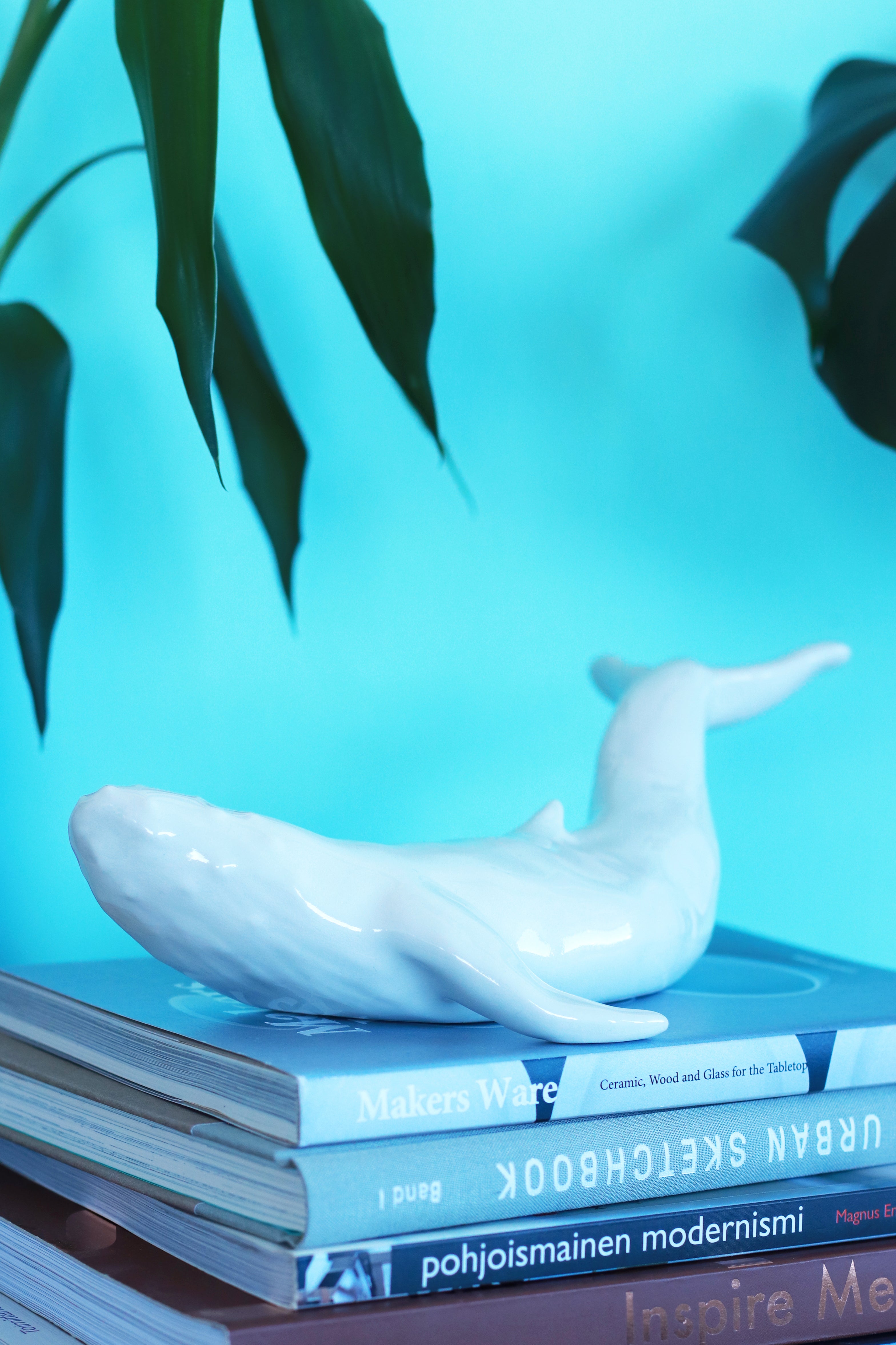 White ceramic humpback whale artwork that is placed on top of art and design books. The background is turquoise and there are plant leaves against it.