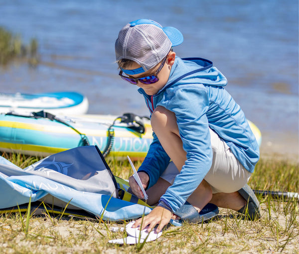 Young boy sitting on grassy spot putting the fins on his NAUTICAL kids standup paddleboard