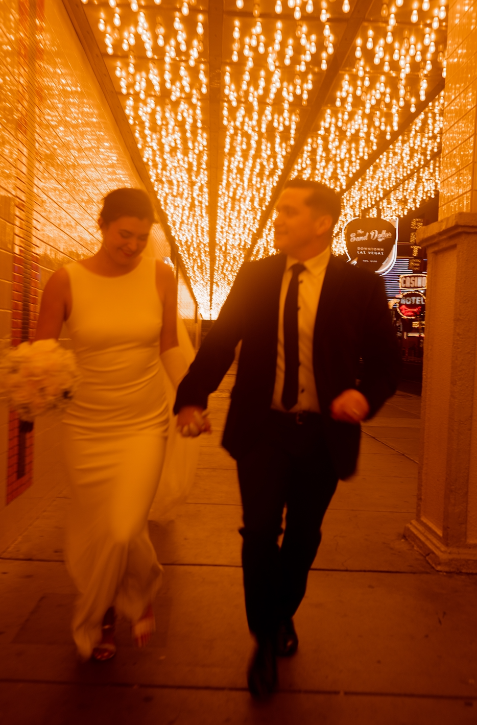 Downtown Las Vegas newlywed couple, running through DTLV with glittery lights in the background. The bride is wearing a perfectly altered gown and the groom a perfectly tailored black suit.