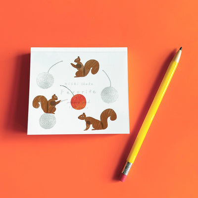 small memo pad illustrated with squirrels on a bright orange background. A yellow pencils lies on the left. 