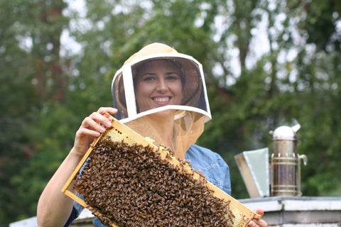 Jodie in beekeeping helmet holding a frame of bees, hive and smoker in background.